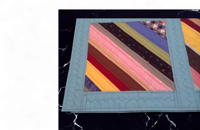 Op de afbeelding: Een handgemaakte, gewatteerde placemat met een lichtblauwe rand. Het midden heeft diagonale strepen in verschillende kleuren, waaronder roze, geel en blauw. De rand heeft een decoratieve, golvende steek.