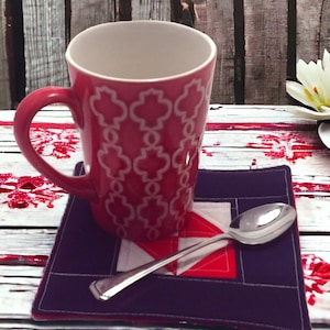 Puede incluir: Un posavasos acolchado morado con un diseño geométrico blanco y rojo. Una taza rosa con un patrón geométrico blanco se encuentra sobre el posavasos. Una cuchara de plata se coloca sobre el posavasos.