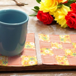 Könnte beinhalten: Zwei orangefarbene und gelbe Stoffuntersetzer mit Blumenmuster. Eine blaue Keramiktasse steht auf einem der Untersetzer.