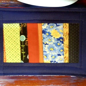 Puede incluir: Un salvamanteles acolchado azul marino con un diseño de patchwork. El patchwork está hecho de tiras de tela en varios colores y patrones, incluyendo verde, naranja, marrón, amarillo y azul.