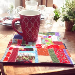 May include: A red mug with a white geometric pattern sits on a colorful patchwork quilt coaster. The coaster is made of various fabric scraps sewn together.