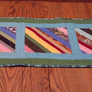 Op de afbeelding: Een rechthoekige, handgemaakte tafelloper met een patchwork-ontwerp. De loper heeft drie centrale rechthoekige panelen met diagonale strepen in verschillende kleuren, omlijst door een lichtblauwe rand en een groene buitenrand. Geplaatst op een houten tafel.