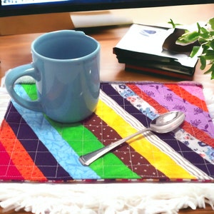 Puede incluir: Un posavasos de tela a rayas de colores con una taza azul claro y una cuchara de plata en la parte superior. El posavasos está sobre una superficie blanca y esponjosa.