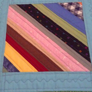 Op de afbeelding: Close-up van een kleurrijke quilt met een lichtblauwe rand. De quilt heeft diagonale strepen in verschillende kleuren, waaronder roze, bruin, blauw en geel. De rand heeft een decoratieve, golvende steek.