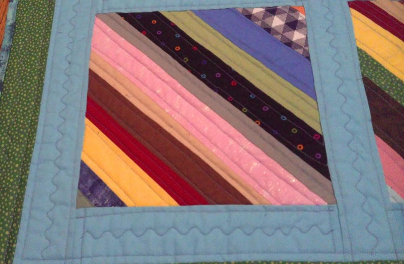 Op de afbeelding: Close-up van een kleurrijke quilt met een lichtblauwe rand. De quilt heeft diagonale strepen in verschillende kleuren, waaronder roze, bruin, blauw en geel. De rand heeft een decoratieve, golvende steek.