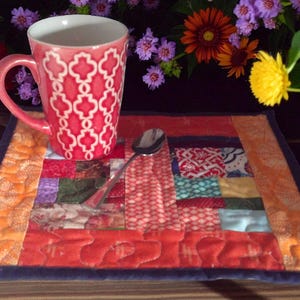 Puede incluir: Una taza rosa con un patrón geométrico blanco se encuentra sobre un colorido mantel individual acolchado. Una cuchara plateada descansa sobre el mantel individual. El fondo presenta flores vibrantes en rojo, amarillo y morado.