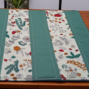 May include: A rectangular teal table runner with two cream-colored floral patterned panels. The floral design includes red, orange, and green accents. The runner is on a wooden table, with sunflowers in vases on either side.