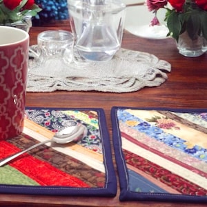 May include: Two colorful fabric placemats with a diagonal striped pattern. A red mug with a white geometric design sits on one of the placemats. A silver spoon is on the placemat next to the mug.
