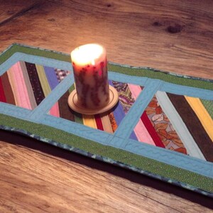 Op de afbeelding: Een brandende kaars op een houten onderzetter staat op een handgemaakte tafelloper. De loper heeft een patchwork-ontwerp met blauwe en groene randen en stroken stof in roze, bruin en geel. De kaars is een decoratief element.