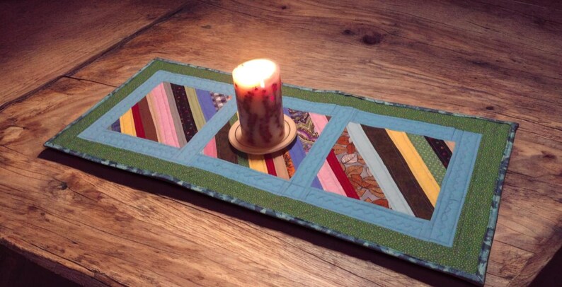 Op de afbeelding: Een brandende kaars op een houten onderzetter staat op een handgemaakte tafelloper. De loper heeft een patchwork-ontwerp met blauwe en groene randen en stroken stof in roze, bruin en geel. De kaars is een decoratief element.