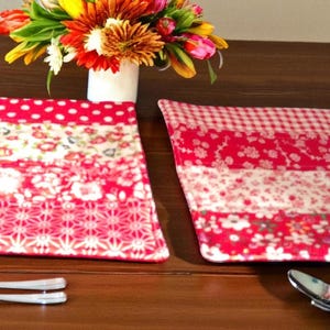 Puede incluir: Dos manteles individuales estampados en rojo y blanco con diseños florales y de lunares están colocados sobre una mesa de madera oscura. La cubertería, incluidos tenedores y cucharas, está dispuesta alrededor de los manteles individuales. Un jarrón con flores coloridas está en el fondo.