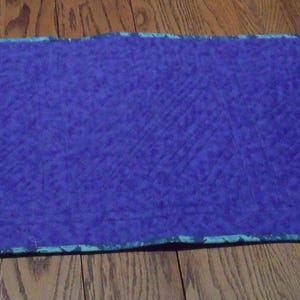 Op de afbeelding: Een rechthoekige, gewatteerde placemat in een diepblauwe tint. De randen zijn afgewerkt met een patroonrand in tinten van blauwgroen en blauw. De placemat ligt op een houten oppervlak.