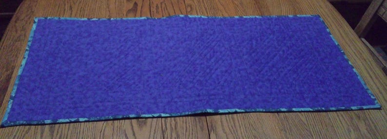 Op de afbeelding: Een rechthoekige, gewatteerde placemat in een diepblauwe tint. De randen zijn afgewerkt met een patroonrand in tinten van blauwgroen en blauw. De placemat ligt op een houten oppervlak.