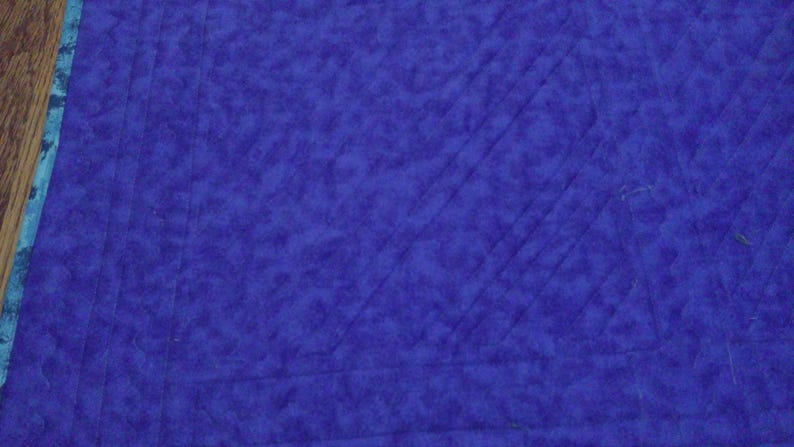 Op de afbeelding: Close-up van een gewatteerde stof in een diepblauwe tint. De stof heeft een getextureerde uitstraling met diagonale stiksels die een ruitpatroon cre&euml;ren. Een blauwgroene en blauwe patroonrand is zichtbaar aan de linkerkant.