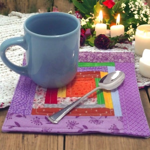 May include: A purple quilted coaster with a colorful patchwork design. A blue mug and a silver spoon are on top of the coaster. Three white candles are in the background.