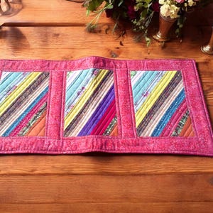 May include: A rectangular, handmade table runner with a pink border. The center features three colorful, angled patchwork panels with various fabrics in shades of blue, yellow, red, and green. The runner is placed on a wooden table.