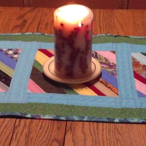 Op de afbeelding: Een handgemaakte tafelloper met een patchwork-ontwerp met diagonale strepen in verschillende kleuren. De loper heeft een lichtblauwe rand en een groene buitenrand. Er staat een brandende kaars op de loper.