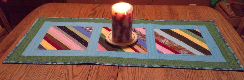 Op de afbeelding: Een handgemaakte tafelloper met een patchwork-ontwerp met diagonale strepen in verschillende kleuren. De loper heeft een lichtblauwe rand en een groene buitenrand. Er staat een brandende kaars op de loper.