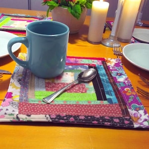 May include: A teal mug sits on a colorful, patchwork placemat with a spoon. The placemat features a variety of fabrics in pink, green, and blue. White plates and silverware are also on the wooden table, along with candles and a floral arrangement.