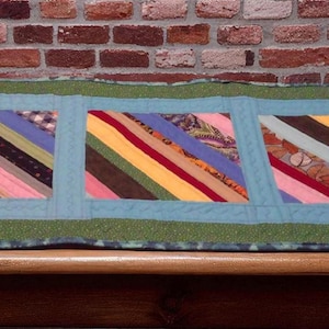 Könnte beinhalten: Handgefertigter Tischläufer mit drei farbenfrohen rechteckigen Paneelen. Jedes Paneel zeigt diagonale Streifen in verschiedenen Farben, darunter Rosa, Blau, Gelb und Braun. Die Paneele sind von einem hellblauen Rand und einem grünen Außenrand umgeben.