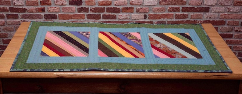 Op de afbeelding: Handgemaakte tafelloper met drie kleurrijke rechthoekige panelen. Elk paneel heeft diagonale strepen in verschillende kleuren, waaronder roze, blauw, geel en bruin. De panelen zijn omlijst door een lichtblauwe rand en een groene buitenrand.