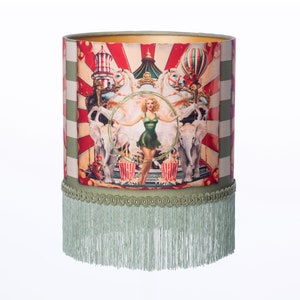 May include: Cylindrical lampshade with a circus design. The shade has a vintage illustration of a woman in a green outfit, elephants, carousel horses, and popcorn. The top and bottom edges are trimmed with green fabric and fringe.