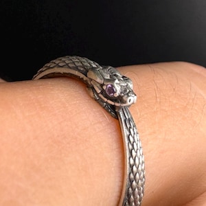 May include: A silver snake ring with a detailed head and textured body. The snake's eye is a small, purple gemstone. The ring is worn on a wrist, showcasing the intricate design and craftsmanship.