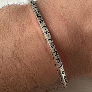 Può includere: Un braccialetto rigido in argento con un'iscrizione nera di rune attorno alla fascia.