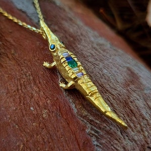 May include: A gold necklace with a detailed crocodile pendant featuring colourful gemstones. The pendant is set against a rustic wooden background.
