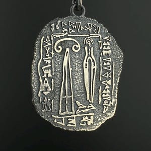 Puede incluir: Un colgante de tono plateado en una cadena delicada, con una forma irregular y texturizada similar a una moneda. La superficie muestra símbolos grabados y dos figuras centrales, una que se asemeja a una forma alta y delgada y la otra a una figura estilizada y abstracta.