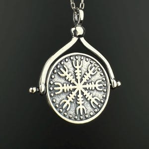 May include: A silver-toned pendant necklace featuring a circular medallion suspended from a rotating frame. The medallion displays a raised Vegvisir symbol, also known as a Viking compass, surrounded by a beaded border on a textured metallic surface.