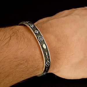 May include: A silver bracelet with a black band featuring a series of symbols and designs.