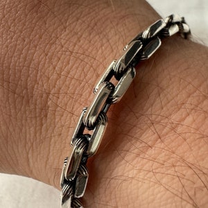 Könnte beinhalten: Ein silbernes Kettenarmband mit einem dicken, seilähnlichen Design. Das Armband besteht aus ineinandergreifenden Gliedern, die flach und rechteckig sind.