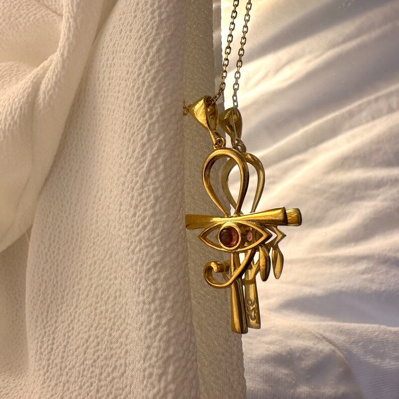 Op de afbeelding: Een gouden ketting met een hanger in de vorm van een ankh met een roze oog in het midden. De ankh is een symbool van leven en onsterfelijkheid in de oude Egyptische cultuur.
