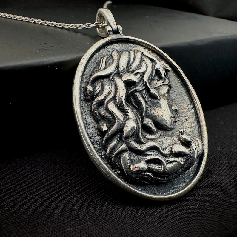 May include: A silver pendant necklace featuring a detailed relief of Medusa's head with snakes for hair. The pendant is oval shaped and hangs from a silver chain.