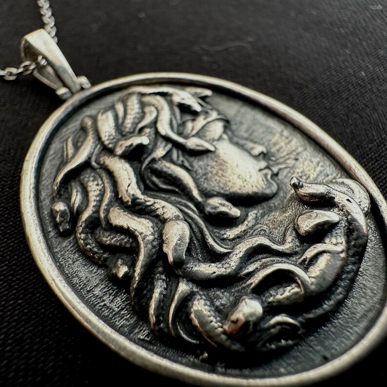 May include: A silver pendant featuring a detailed relief sculpture of a woman's face with snakes in her hair. The pendant is oval shaped and has a textured surface.