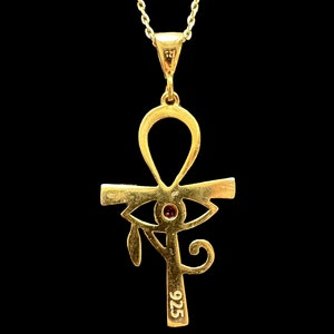 Op de afbeelding: Een gouden ketting met een hanger in de vorm van een ankh-symbool met een rood oog in het midden. De hanger is aan de achterkant gemarkeerd met "925".
