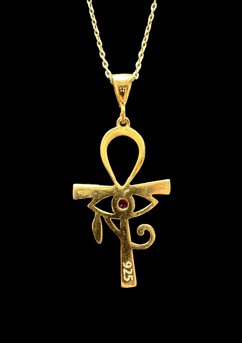 Op de afbeelding: Een gouden ketting met een hanger in de vorm van een ankh-symbool met een rood oog in het midden. De hanger is aan de achterkant gemarkeerd met "925".
