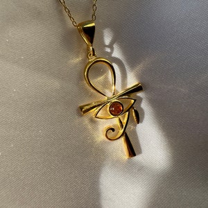 Op de afbeelding: Een gouden ketting met een hanger in de vorm van een ankh-symbool met een oranje oog van Horus. De hanger hangt aan een delicate gouden ketting.