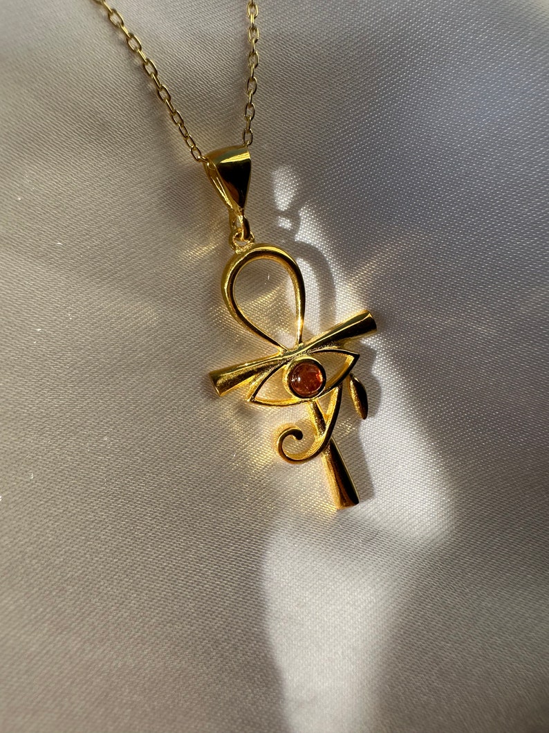 Op de afbeelding: Een gouden ketting met een hanger in de vorm van een ankh-symbool met een oranje oog van Horus. De hanger hangt aan een delicate gouden ketting.