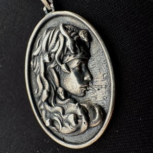 May include: Silver pendant featuring a detailed relief of a woman's face with a snake in her hair. The pendant is round and has a smooth, polished surface.