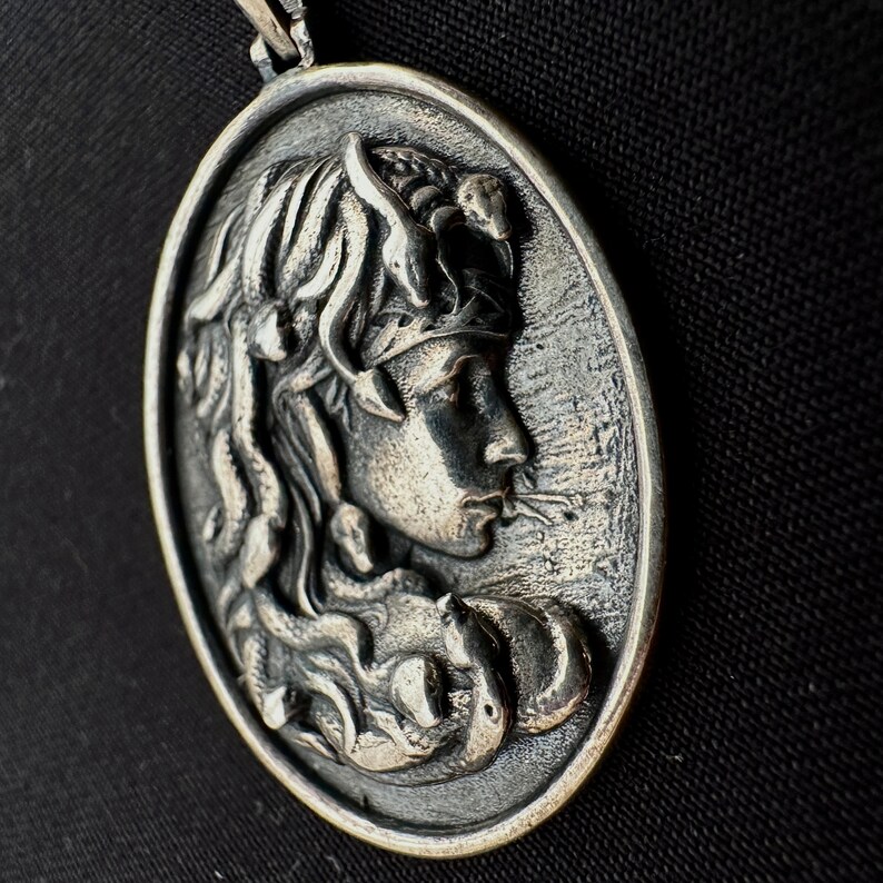May include: Silver pendant featuring a detailed relief of a woman's face with a snake in her hair. The pendant is round and has a smooth, polished surface.