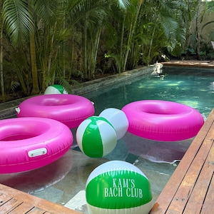 May include: A swimming pool scene with pink inflatable rings and green and white beach balls floating in the water. The text "KAM'S BACH CLUB" is visible on one of the beach balls. Lush green palm trees surround the pool.