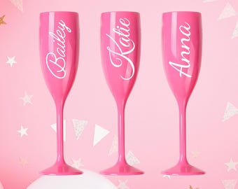 May include: Three pink champagne flutes with white lettering. The names "Bailey", "Katie", and "Anna" are written on the flutes. The background is pink with white and gold star and triangle confetti.