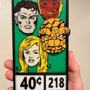 May include: A rectangular, green and black framed comic book art piece featuring four character portraits. The portraits include a man with brown hair, a flaming figure, a rocky-skinned character, and a blonde woman. The bottom section displays the text "40¢" and "218 MAY 02462".