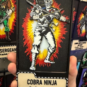 May include: A black framed collectible featuring a Cobra Ninja character, Storm Shadow, from the G.I. Joe franchise. The figure is depicted in a dynamic pose with two swords against a red, yellow, and orange burst background. The text "COBRA NINJA" and "STORM SHADOW" are displayed.