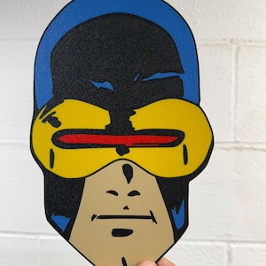May include: A flat, die-cut graphic of a comic book character head featuring a blue hood, a black mask, a bright yellow visor, and a thin horizontal red beam across the center. The character has a tan chin and jawline with black line details.