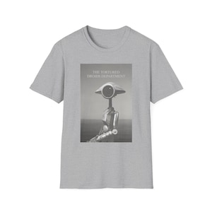 May include: A light gray t-shirt featuring a black and white image of a robot with the text "THE TORTURED DROIDS DEPARTMENT". The robot has a large, round head and a slender body. The shirt is made of a soft material.