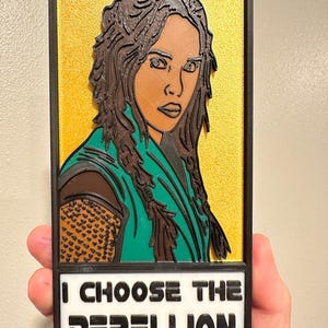 May include: A 3D-printed portrait of a woman with brown hair and green clothing, set against a gold background. The image is framed in black and features the text "I CHOOSE THE REBELLION" in bold white letters.