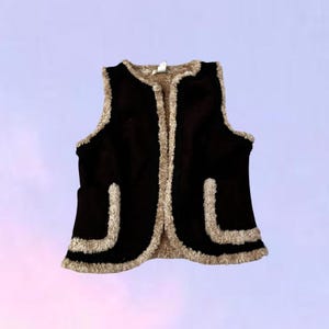 May include: A sleeveless vest in dark brown with a soft, beige faux-shearling trim along the edges, neckline, and two front pockets. The garment features a smooth, dark brown exterior and a plush, light-colored interior lining visible at the seams.